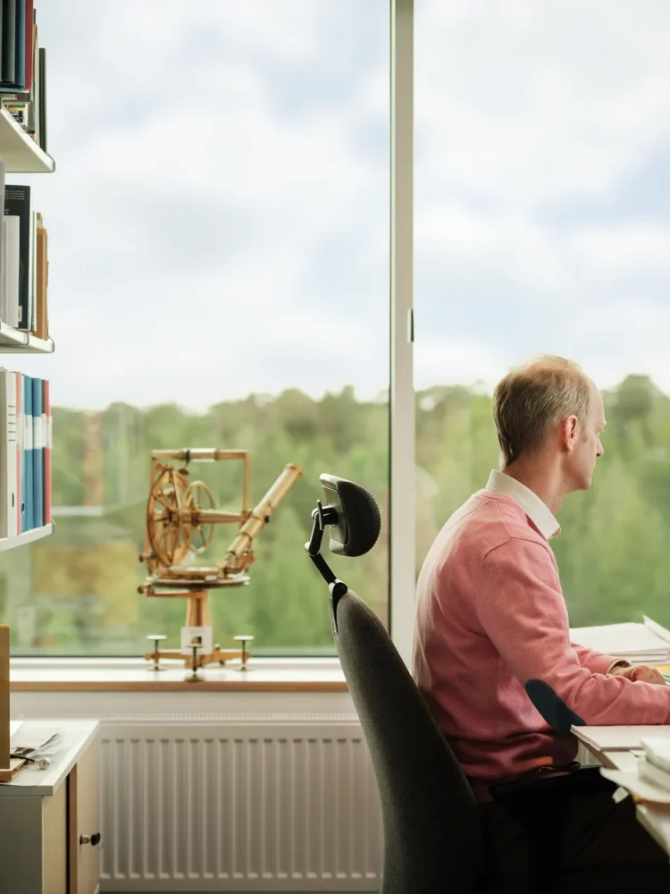 Man in pink sweater sitting at desk with large windows, surrounded by books and scientific instruments.