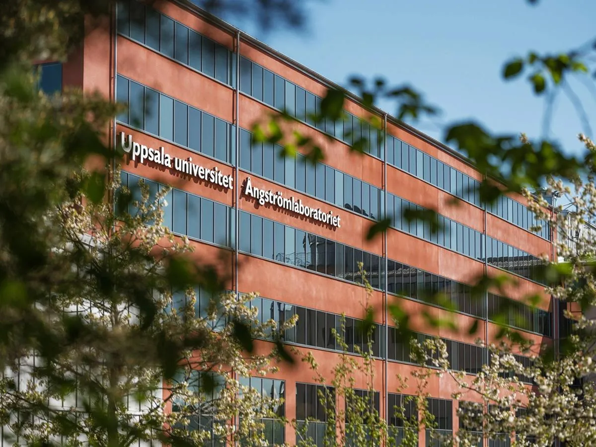Bâtiment rouge comportant de grandes baies vitrées et portant l'inscription « Uppsala University Ångström Laboratory » sur la façade, partiellement masqué par des arbres.