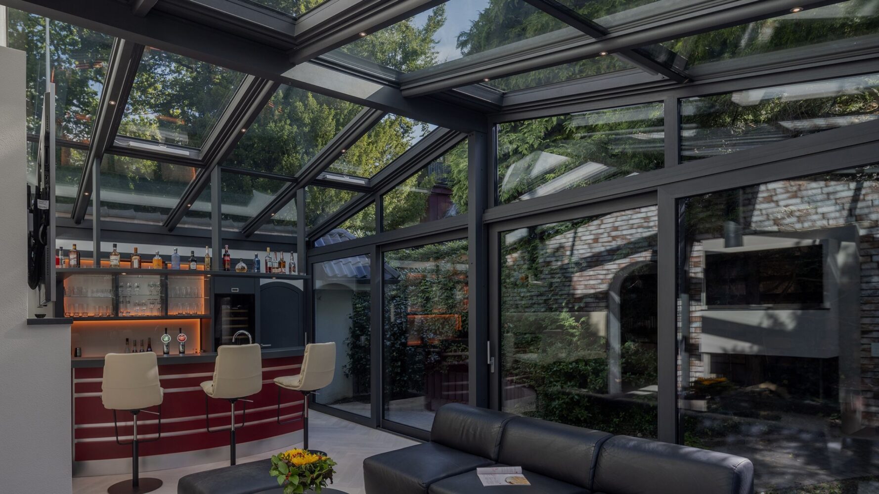 Glass pavilion in a garden setting with a black frame and open glass sections that let in daylight.