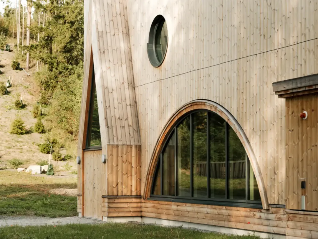 Close-up of a wood-clad building with rounded windows and vertical panels in light tones, surrounded by greenery.