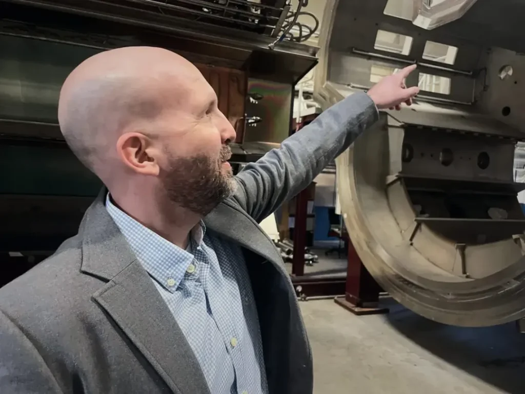Porträt eines Mannes in einer grauen Jacke, der in einer Industrieumgebung steht, im Hintergrund sind große Maschinenteile zu sehen.