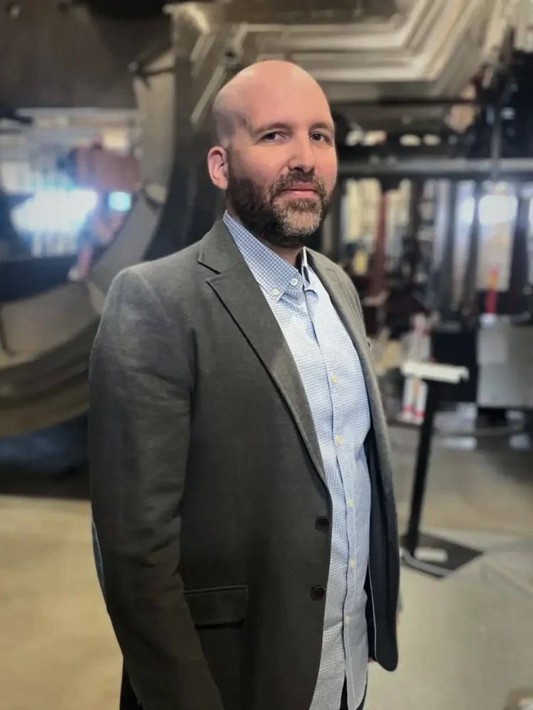 Portrait of a man in a gray jacket standing in an industrial environment with large machine parts in the background.