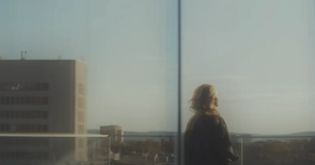 Reflected image of a woman in a glass facade, with doubled contours and a background of sky and high-rise buildings.