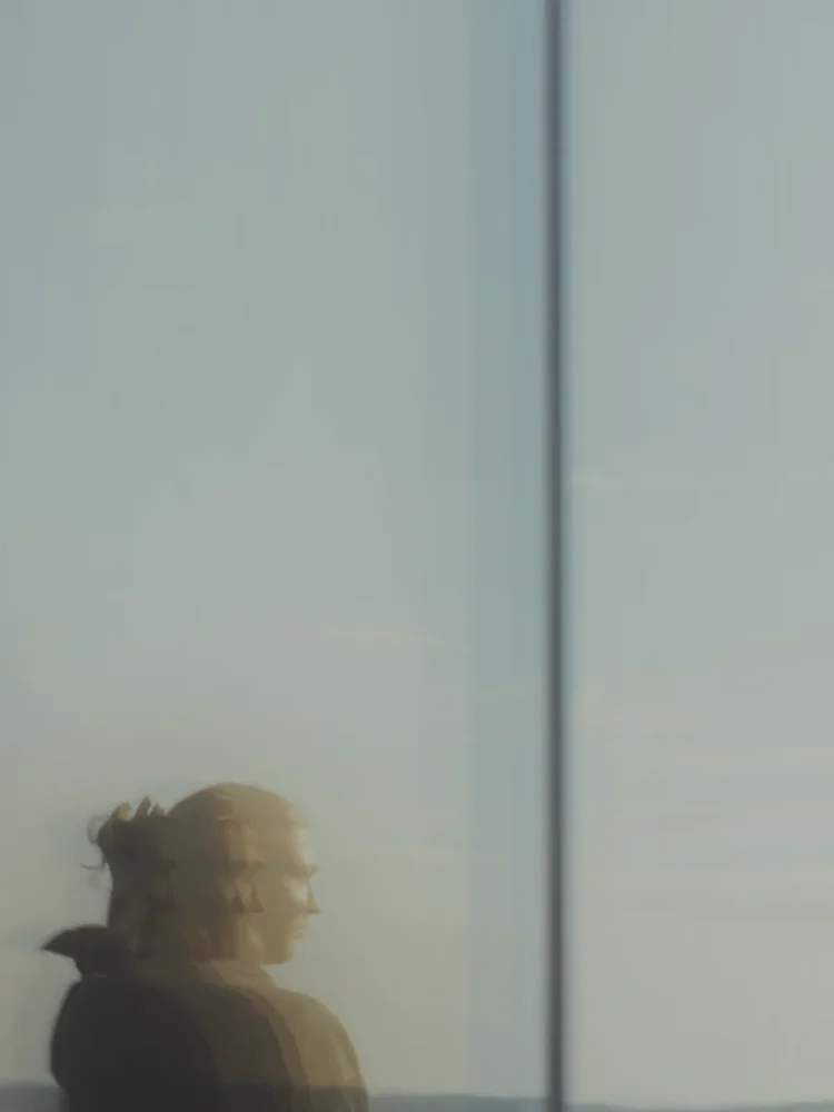 Reflected image of a woman in a glass facade, with doubled contours and a background of sky and high-rise buildings.