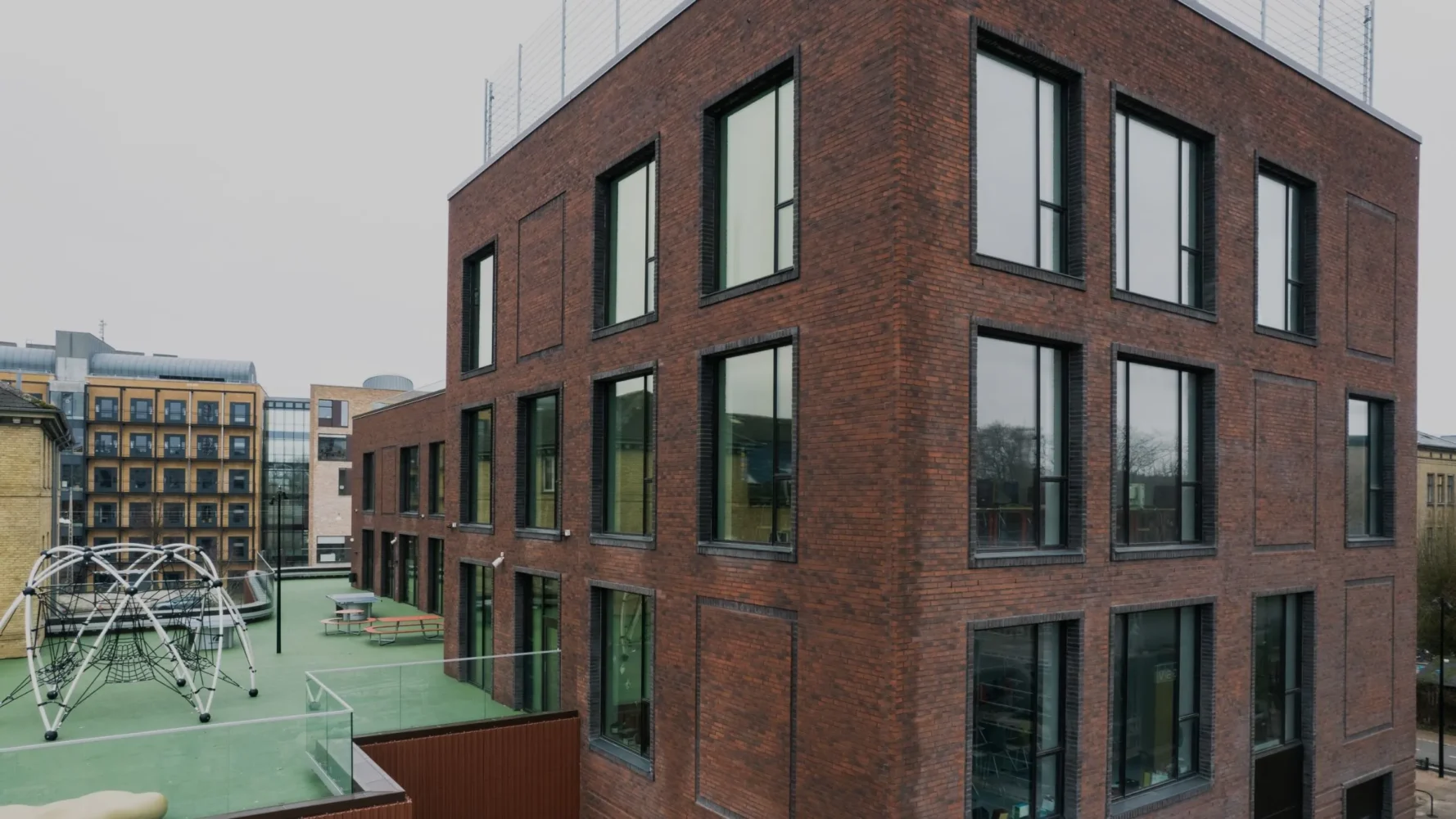 Modern skolbyggnad i mörkrött tegel med stora fönster och grönt utomhusgolv, fotograferad i vinkel.