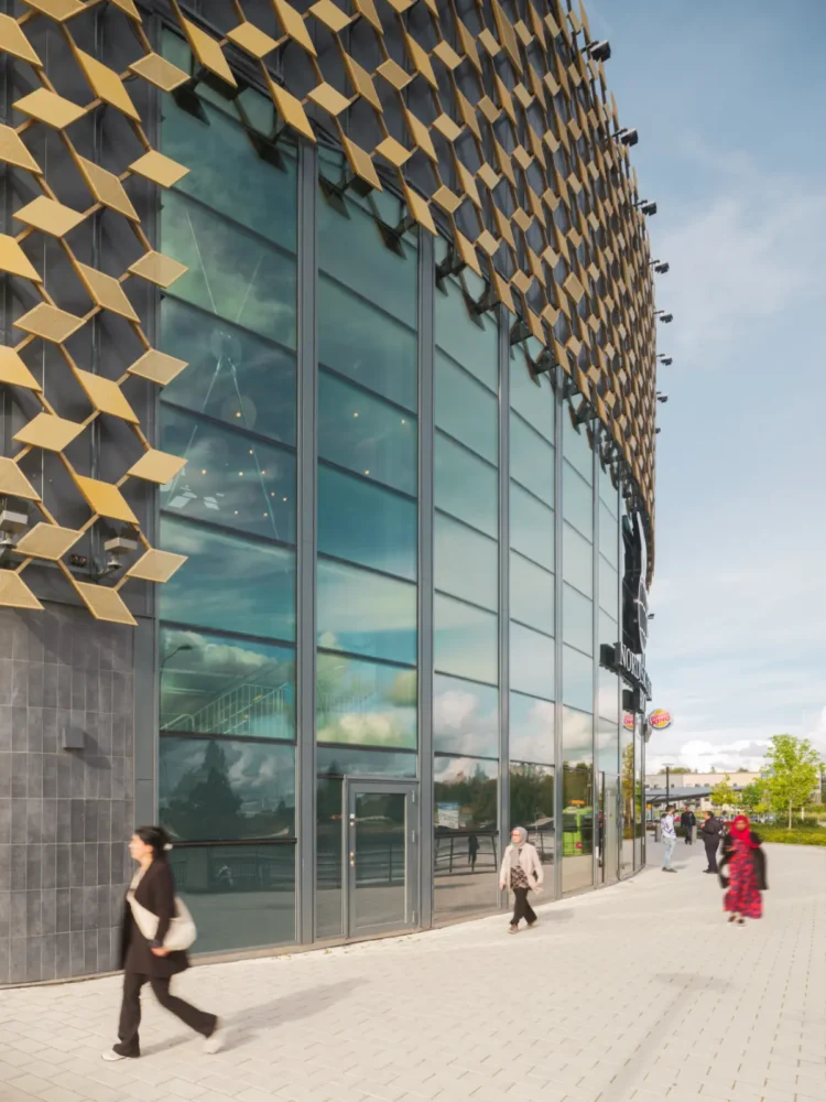 Curved glass facade of a building with gold-colored facade elements, people walking on the sidewalk in front.