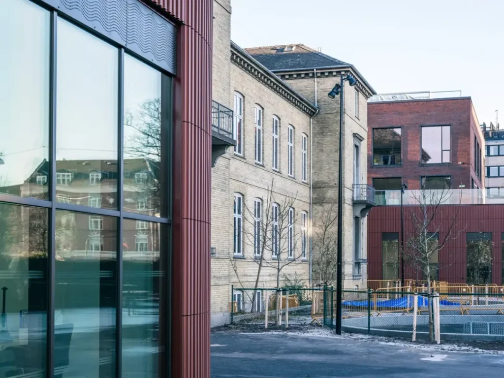Exterior view of an older school building with beige brick, modern extensions and large glass panels in different color tones.