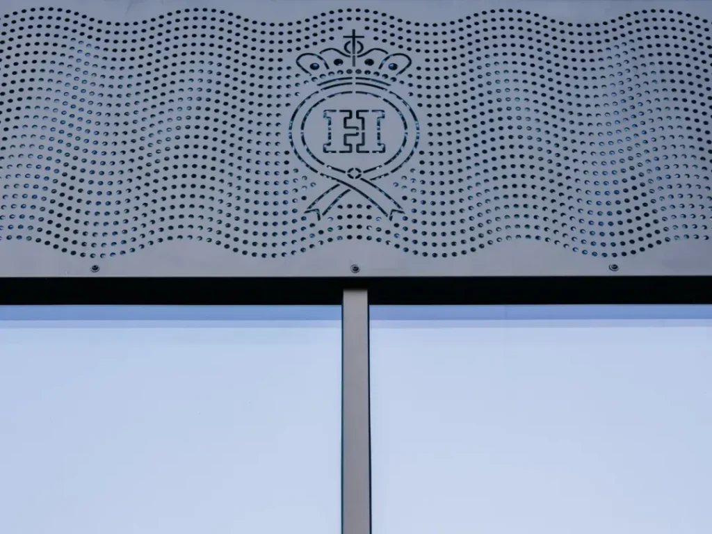 Detaljbild på perforerad metallfasad med ett dekorativt mönster och bokstaven "H&" i mitten.