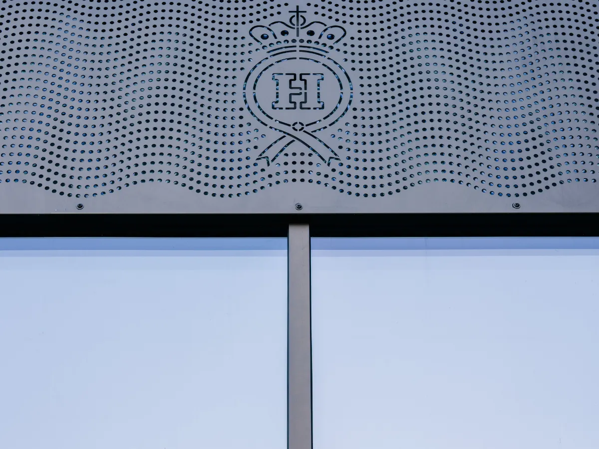 Gros plan sur un panneau métallique perforé situé sur la façade d'un bâtiment, orné d'un motif décoratif ondulé et d'un emblème en forme de blason avec la lettre « H » au centre, situé au-dessus d'un châssis de fenêtre vertical.